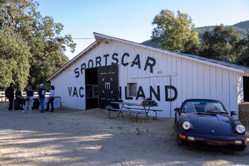 A classic black Porsche is parked in front of a white barn labeled "Sportscar Vacilland," drawing a crowd during Car Week—a perfect off the beaten path spot for your next sportscar vacation.