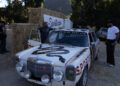 A vintage Mercedes sportscar with custom racing decals is parked by hay bales, as several people gather around it outdoors during Car Week in Vacationland.