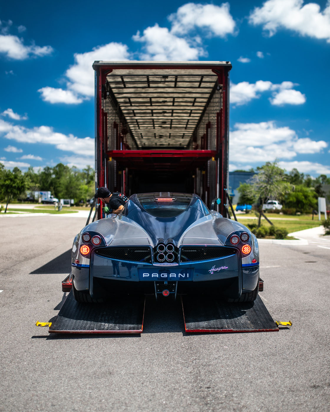 A Pagani Huayra sports car is being loaded into an enclosed transport trailer on a sunny day at St. Armands Circle in Sarasota, with a person assisting at the rear during an exotic car show.