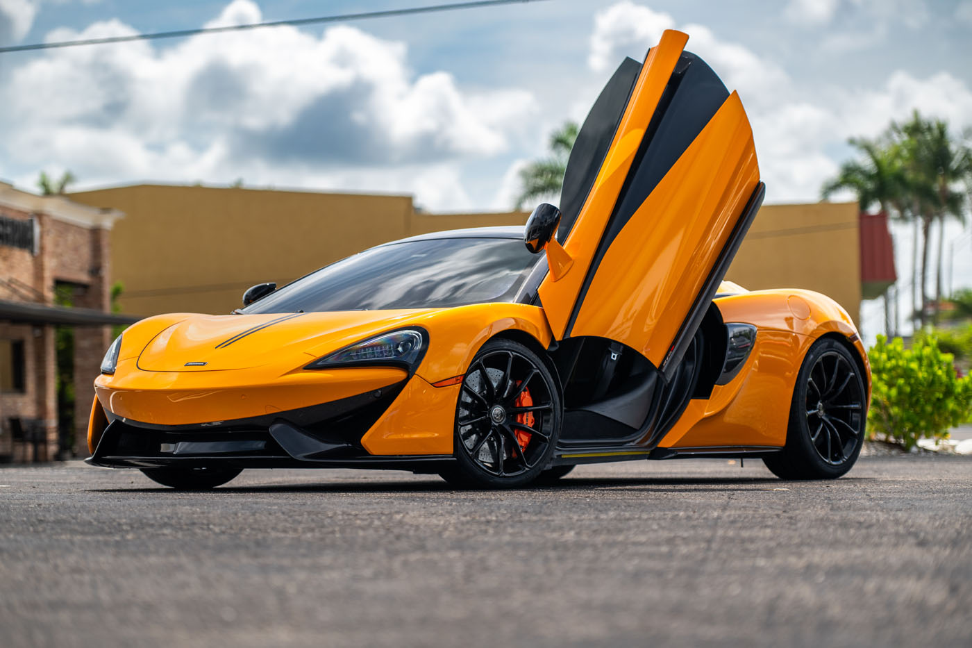 An orange sports car with its driver-side door open upwards is parked outdoors on a sunny day at the St. Armands Circle Exotic Car Show in Sarasota.