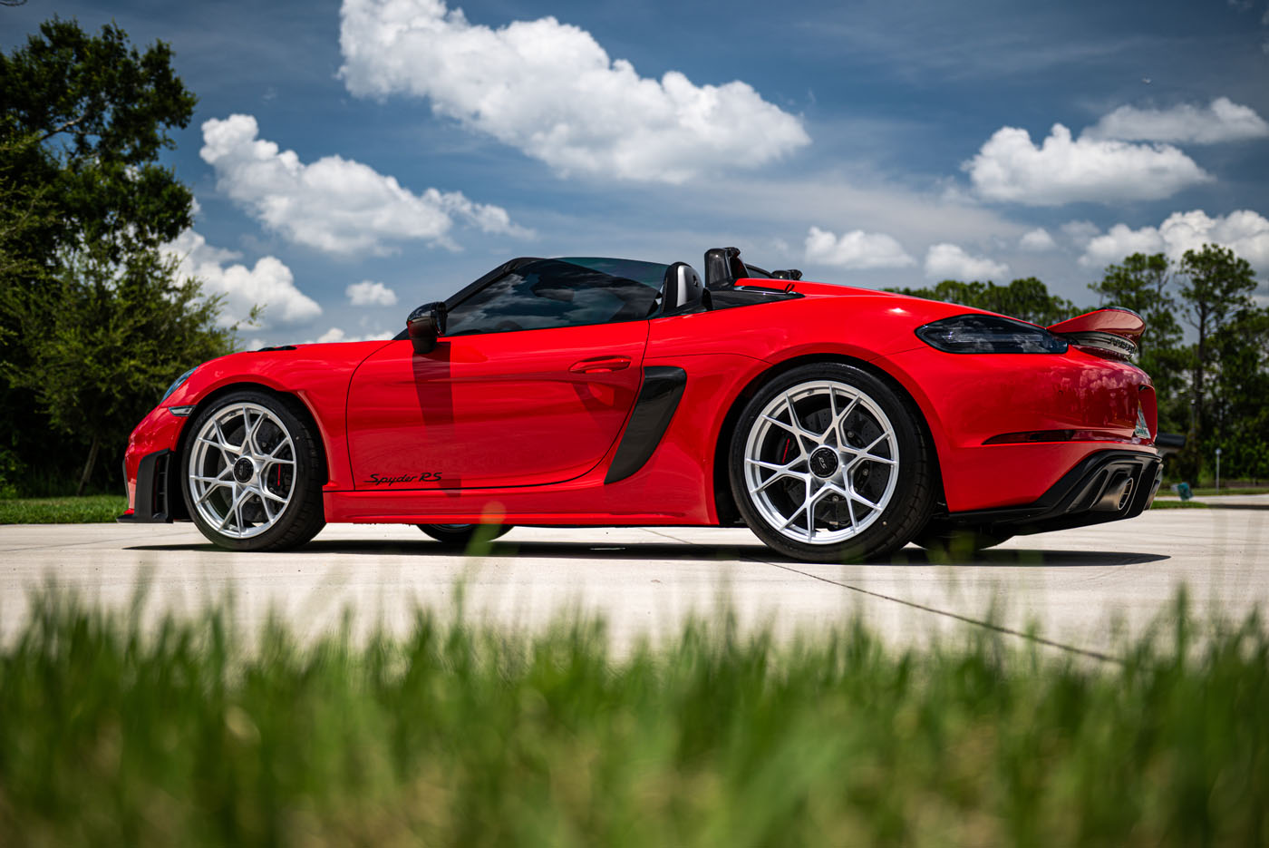 A red convertible sports car parked on a concrete driveway, with green grass in the foreground and trees under a partly cloudy sky, reminiscent of the stunning displays at Sarasota’s Exotic Car Show near St. Armands Circle.