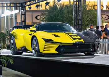 A yellow and black Ferrari Daytona SP3, worth $26 million, is displayed on an outdoor platform at a car show, drawing the attention of billionaire car enthusiasts amid people and lights in the background.