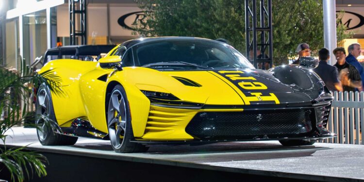 A yellow and black Ferrari Daytona SP3, worth $26 million, is displayed on an outdoor platform at a car show, drawing the attention of billionaire car enthusiasts amid people and lights in the background.