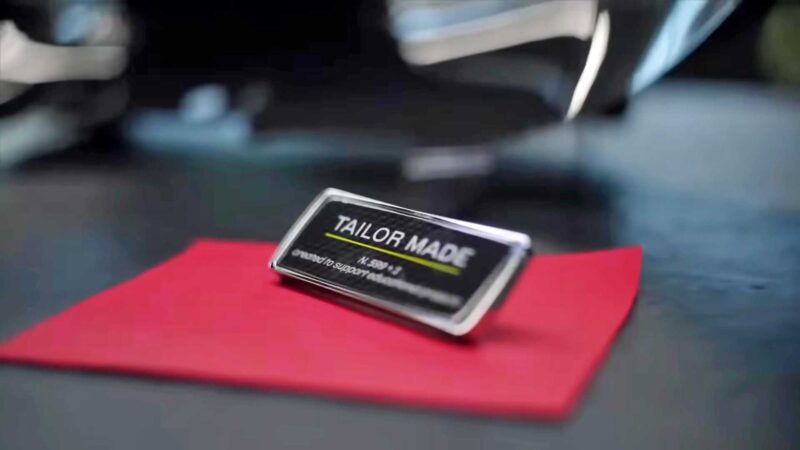 A metallic "Tailor Made" nameplate rests on a red cloth, evoking the exclusivity of a Billionaire's $26 million Ferrari Daytona SP3, with a blurred background.