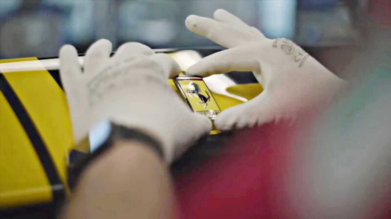 A person wearing white gloves attaches a Ferrari logo badge to a yellow $26 million Ferrari Daytona SP3, embodying the billionaire lifestyle.
