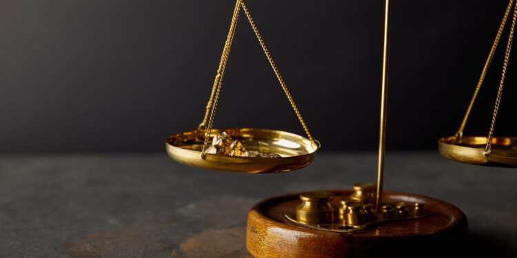 A gold balance scale holds gold nuggets on one side and weights on the other, symbolizing asset valuation, placed on a dark stone surface with a black background.