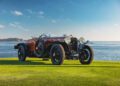 A vintage Hispano-Suiza two-seater convertible is parked on a grassy field by the ocean under a clear blue sky, reminiscent of classic displays at Pebble Beach Concours d’Elegance.