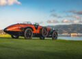 A vintage Hispano-Suiza Tulipwood Torpedo with wood-accented bodywork is parked on grass near a body of water, set against hills and a cloudy sky—a classic moment reminiscent of the Pebble Beach Concours d’Elegance.
