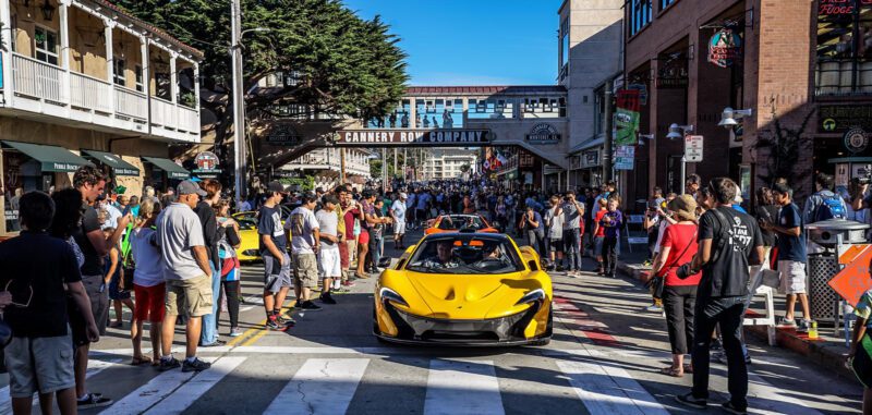 A yellow sports car drives down a crowded street during Monterey Car Week, with shops and a "Cannery Row Company" sign visible in the background—a scene worthy of duPont REGISTRY’s 2025 highlights.