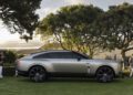 A silver Bentley EXP 15 Concept electric SUV is parked on a grassy area at Pebble Beach, surrounded by trees and people under a partly cloudy sky.