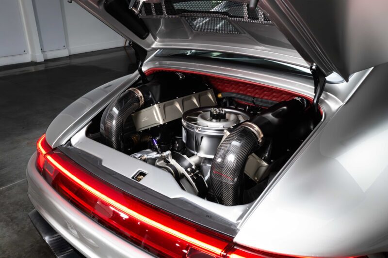 A high-performance sports car with its rear hood open, revealing a detailed view of the engine and carbon fiber components—reminiscent of the Gunther Werks Unveils Limited Edition 1000-HP Slantnose-Inspired F-26 at The Quail—in a well-lit garage.