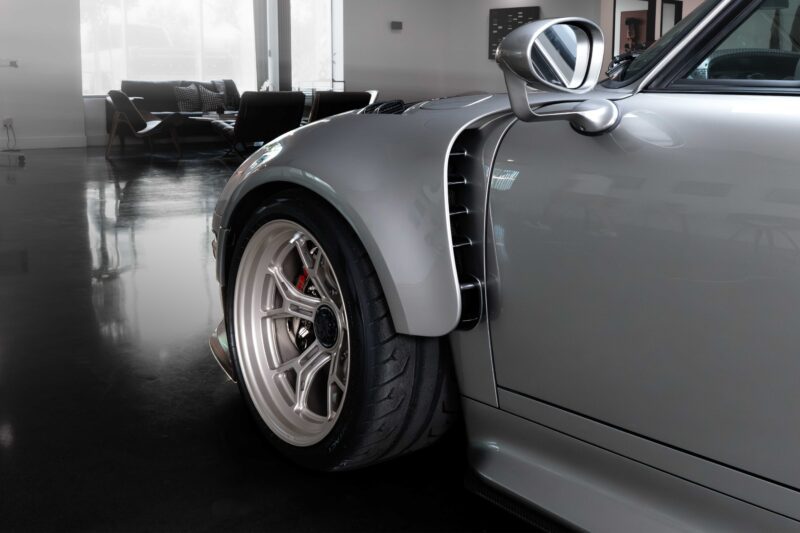 Close-up of the front left side of a silver sports car parked indoors, possibly inspired by the Gunther Werks Unveils Limited Edition 1000-HP Slantnose-Inspired F-26 at The Quail; modern furniture visible in the background.