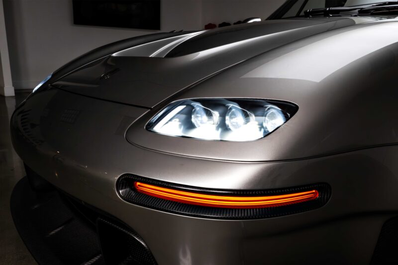 Close-up of the front left headlight and illuminated amber turn signal on a silver sports car, reminiscent of the Gunther Werks Unveils Limited Edition 1000-HP Slantnose-Inspired F-26 at The Quail, in a well-lit indoor setting.