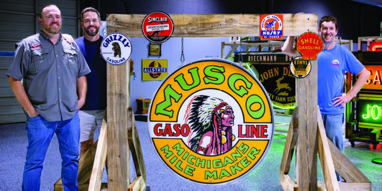 Three men stand beside a large vintage Musgo Gasoline sign, surrounded by collectible signs in an indoor setting—a testament to the enduring investment appeal of retro gas station memorabilia.
