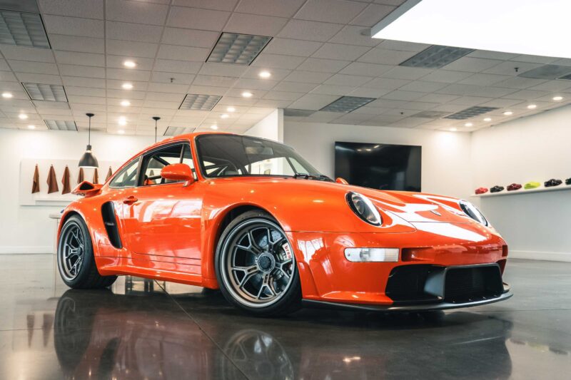 Bright orange Gunther Werks Turbo Tested: Bar None The Gnarliest Porsche Restomod Yet sports car with aerodynamic design and silver rims displayed indoors on a polished floor under ceiling lights.
