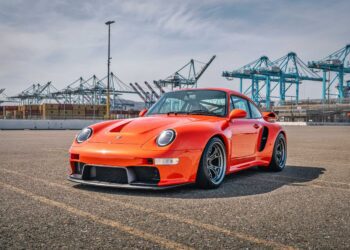 An orange Gunther Werks Turbo Tested sports car is parked on asphalt near a port with shipping containers and cranes in the background under a partly cloudy sky—bar none, the gnarliest Porsche restomod yet.