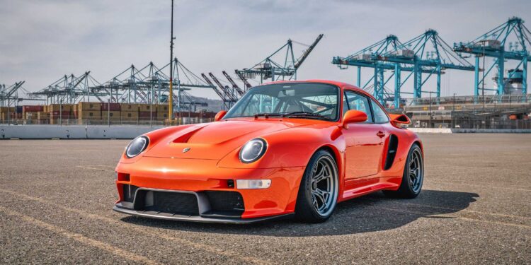 An orange Gunther Werks Turbo Tested sports car is parked on asphalt near a port with shipping containers and cranes in the background under a partly cloudy sky—bar none, the gnarliest Porsche restomod yet.