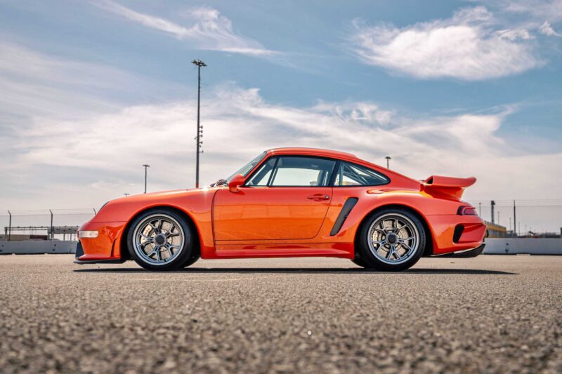 A red Gunther Werks Turbo Tested sports car with a rear spoiler is parked on an empty asphalt lot under a partly cloudy sky, with a fence and light poles in the background—bar none the gnarliest Porsche restomod yet.
