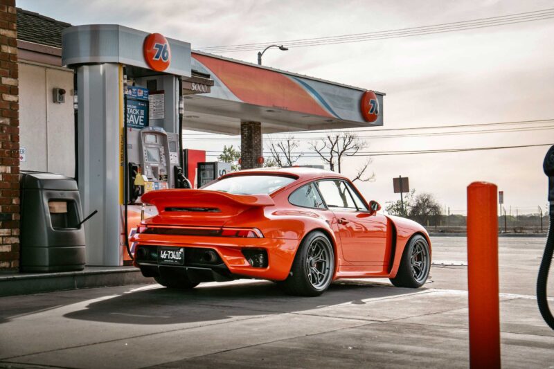 A bright orange Gunther Werks Turbo Tested sports car—the gnarliest Porsche restomod yet—is parked at a 76 gas station pump on a clear day.