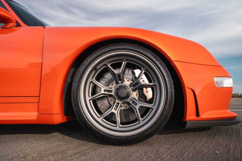 Close-up of the front left wheel and fender of an orange sports car, featuring a black alloy wheel, drilled brake rotor, and red brake caliper—true to the Bar None The Gnarliest Porsche Restomod Yet by Gunther Werks Turbo Tested.