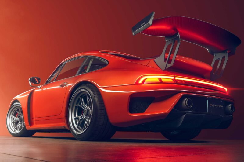 A low-angle view of a Gunther Werks Turbo Tested: Bar None The Gnarliest Porsche Restomod Yet—an orange sports car with a large rear wing, illuminated taillights, and alloy wheels, set against an orange background.