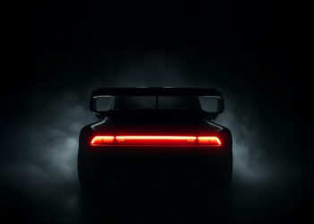 A rear view of a Slantnose-inspired sports car with a large spoiler, illuminated by its red taillight, surrounded by darkness and faint smoke—an unmistakable Gunther Werks presence at The Quail.