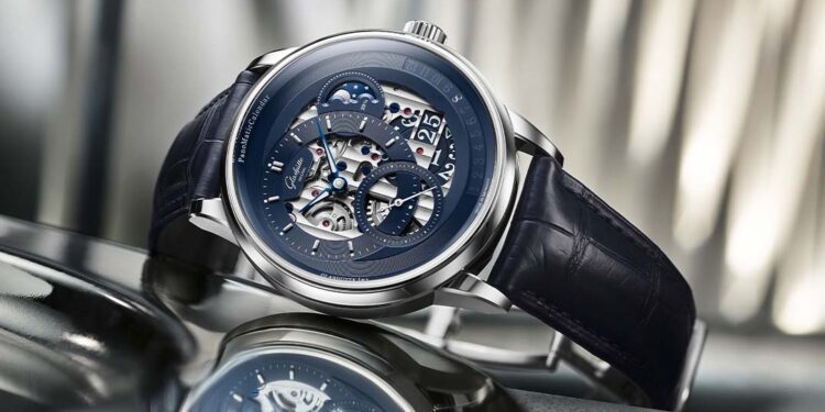 A silver luxury wristwatch by Glashütte Original, featuring a blue skeleton dial and black leather strap, is displayed on a metallic surface with a blurred background—an exquisite limited edition from the PanoMaticCalendar collection.