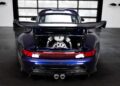 A rear view of a dark blue Gunther Werks Porsche 911 with its engine compartment open, showcasing the remastering of the engine and large rear wing in a modern garage.