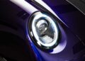 Close-up of a modern car’s round LED headlight with a carbon fiber accent, set against a glossy purple body—an iconic detail in the remastering of the Porsche 911 by Gunther Werks.