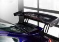 Close-up of a large carbon fiber rear wing spoiler mounted on a dark purple Gunther Werks Porsche 911, positioned indoors near large windows.