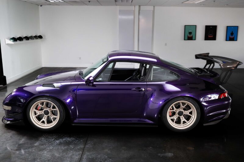 A purple Gunther Werks Porsche 911 with a large rear wing is parked indoors in a minimalist room adorned with framed automotive art on the wall.