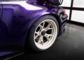 Close-up of the rear wheel and fender of an exclusive purple Gunther Werks GWX sports car, showcasing a white alloy rim and low-profile tire, parked indoors near a window—an impressive Porsche 911 remastering.