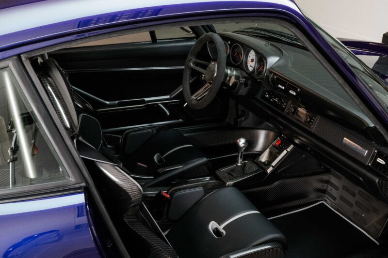 View of a Gunther Werks Porsche 911 interior from the driver’s side, showing two black seats, a manual gear shift, a steering wheel, and a minimalist dashboard with analog gauges—a true remastering of classic design.