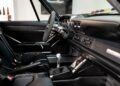 Interior of a remastered Porsche 911 by Gunther Werks, featuring carbon fiber details, a manual gear shift, black bucket seats with harnesses, and a minimalist dashboard.