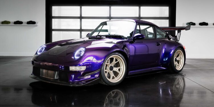 A remastering of the Porsche 911 by Gunther Werks, this purple sports car with a large rear wing is parked indoors on a polished concrete floor, with helmets displayed on shelves in the background.