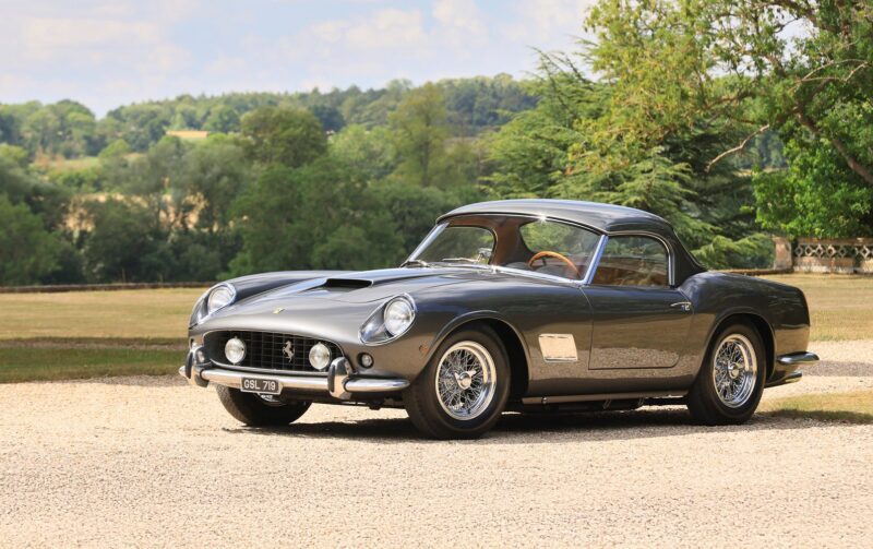 A classic dark gray vintage sports car, featured by duPont REGISTRY, is parked on a gravel driveway with grass, trees, and a blue sky—a timeless scene fit for Monterey Car Week 2025.