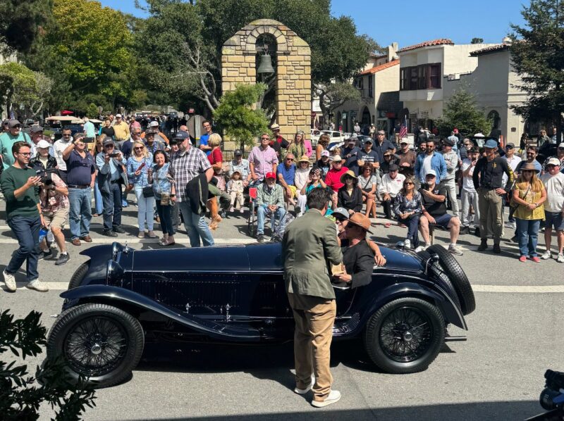 A vintage car is parked on a street during Monterey Car Week as two men talk beside it, surrounded by a large crowd enjoying the 2025 outdoor event.