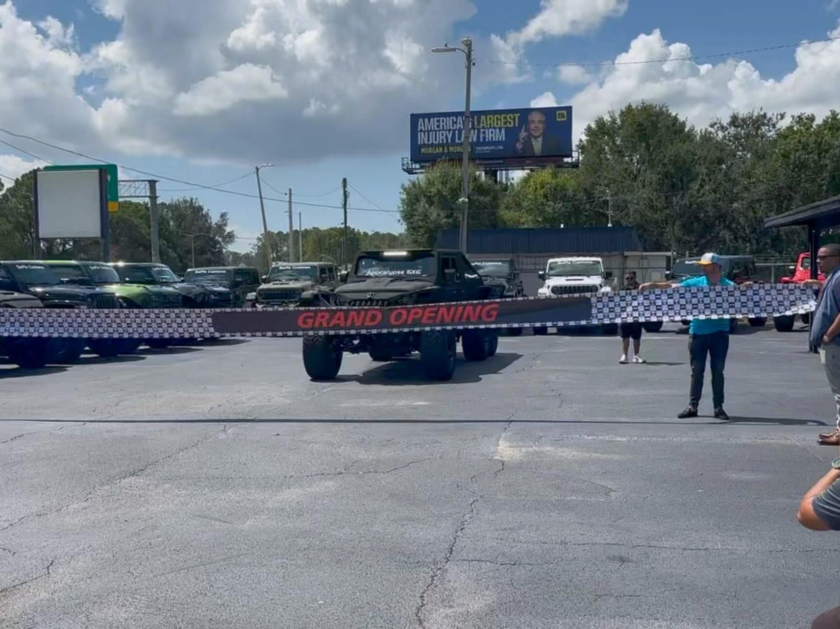 A military vehicle customized by SoFlo Customs bursts through a “Grand Opening Tampa” banner in a parking lot as onlookers watch and photograph the thrilling event under a sunny sky.