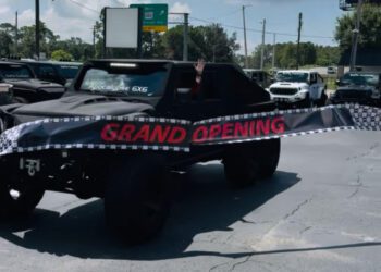 A black 6x6 vehicle from SoFlo Customs drives through a "Grand Opening" banner in a Tampa parking lot, with a person inside waving their hand.