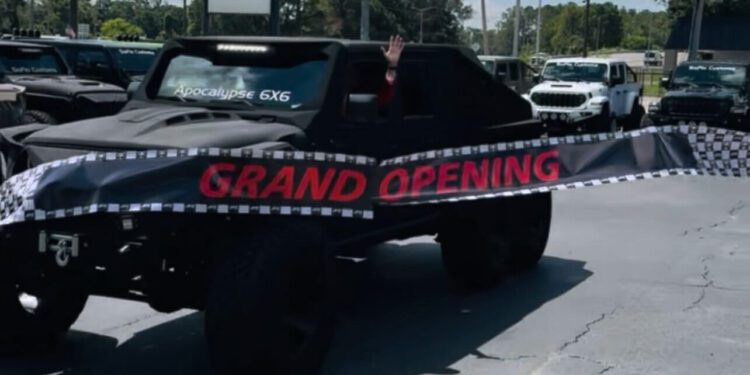 A black 6x6 vehicle from SoFlo Customs drives through a "Grand Opening" banner in a Tampa parking lot, with a person inside waving their hand.
