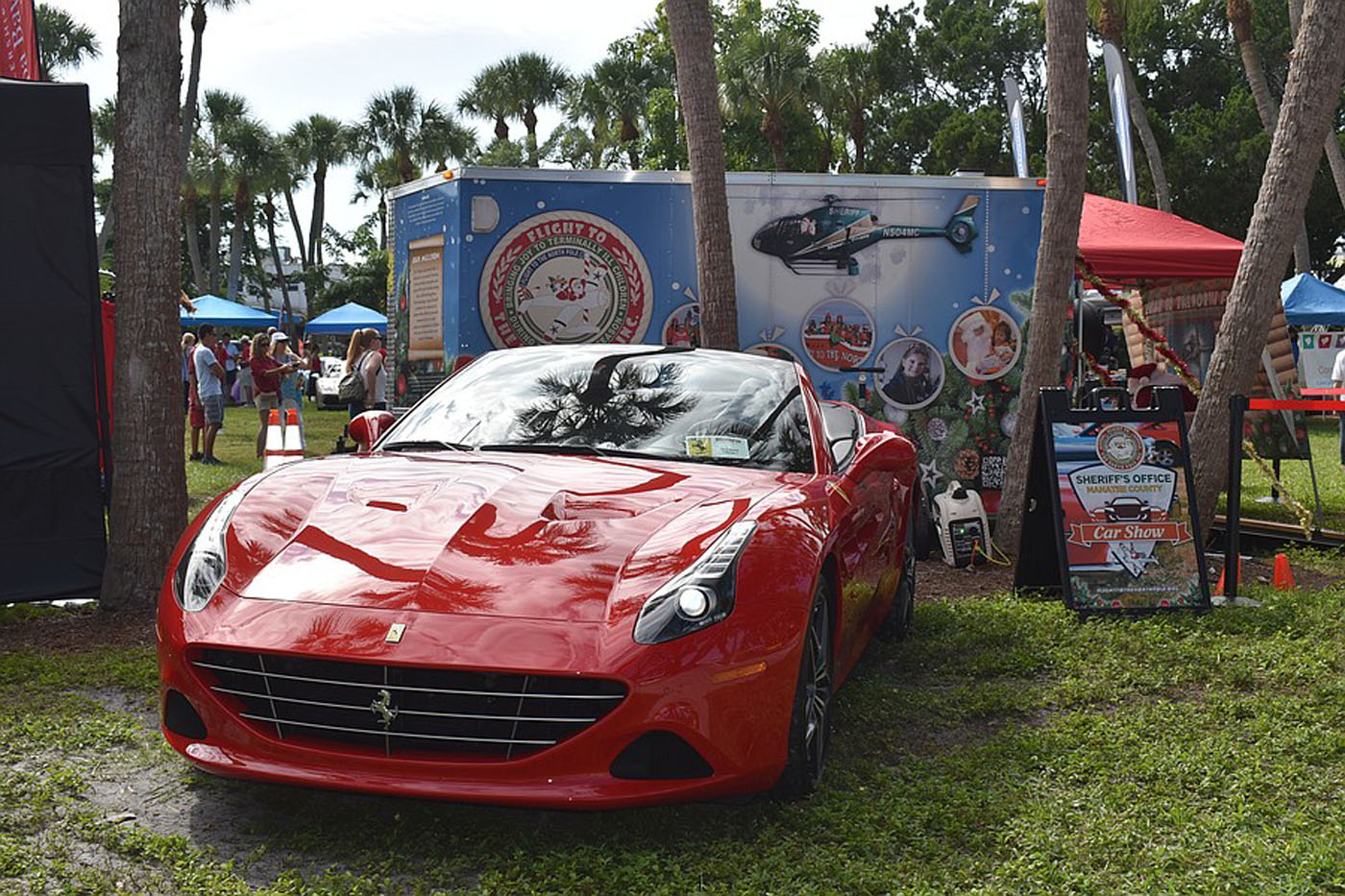 A red Ferrari is parked on grass at an outdoor event at St. Armands Circle, Sarasota, with a Sheriff's Office display and people in the background during the Exotic Car Show.
