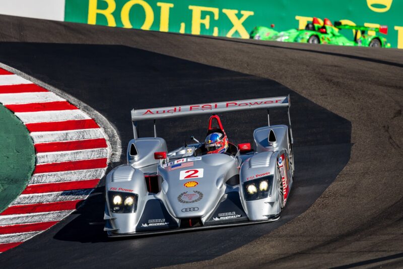 A silver Audi race car with the number 2 speeds around a racetrack curve during Monterey Car Week, with a green race car and a Rolex advertisement in the background.