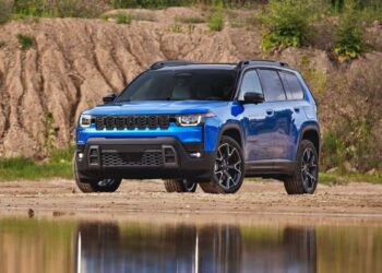 A blue SUV, possibly the 2026 Jeep Cherokee First Look: Rugged Design Hybrid Power and More Space, is parked on a sandy surface near water, with grass and soil mounds in the background.