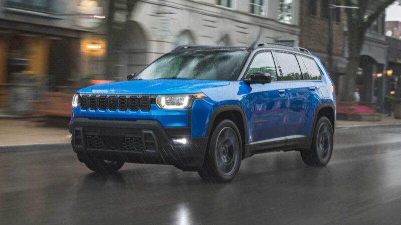 A blue 2026 Jeep Cherokee drives on a wet city street in the rain, showcasing its rugged design and hybrid power, with buildings and trees visible in the background.