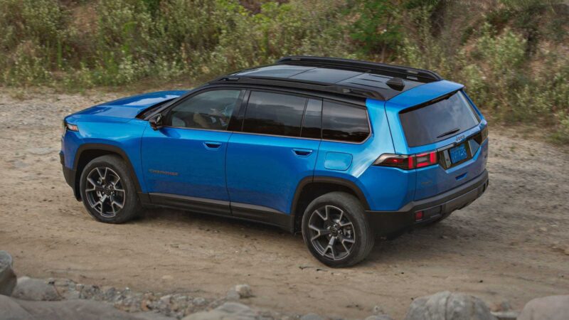 A blue SUV, likely the 2026 Jeep Cherokee, drives on a dirt path surrounded by rocks and greenery—showcasing its rugged design and hinting at hybrid power and more space from a rear-side angle.