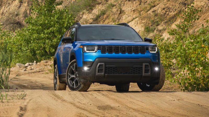 A blue SUV, possibly the 2026 Jeep Cherokee with its rugged design and hybrid power, is parked on a dirt road, surrounded by green bushes and a rocky hillside in the background.