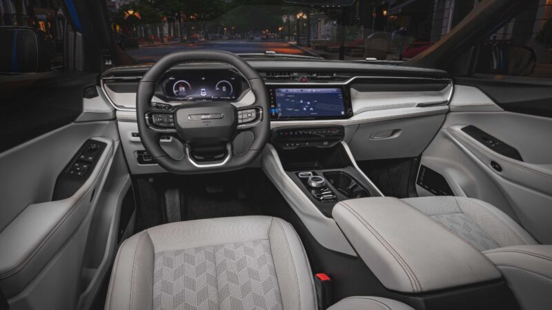 View of a modern car interior featuring a digital instrument cluster, touchscreen display, multifunction steering wheel, light grey seats, and various controls—offering a first look at the 2026 Jeep Cherokee’s rugged design and spacious hybrid-powered cabin.