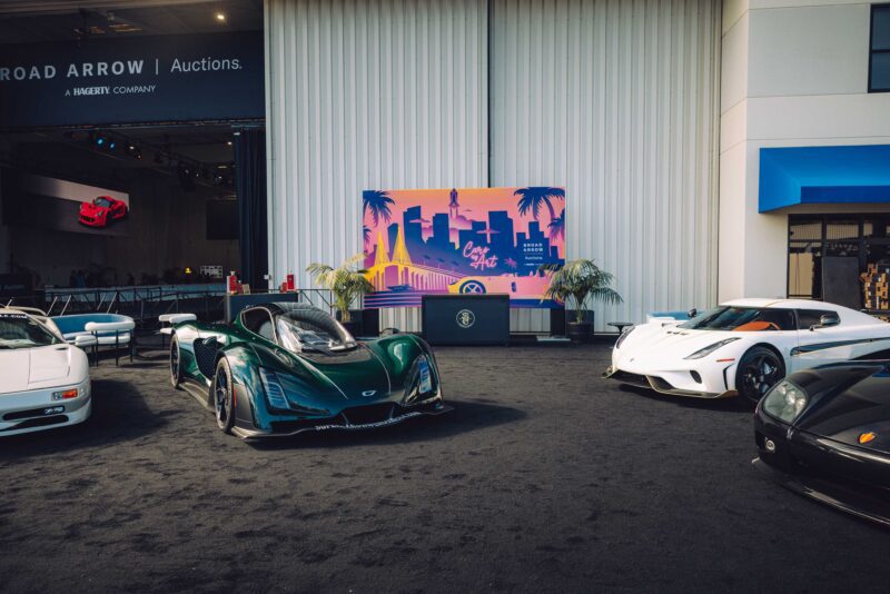 A row of luxury sports cars parked in front of a building with an “East Coast” neon sign and palm graphics sets the stage for SOC’s Monterey Debut, featuring Convoys Exclusives and Touring’s Veloce12 Barchetta Reveal.