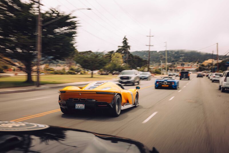 A yellow sports car drives down a multi-lane road alongside other vehicles on an overcast day, echoing the excitement of Touring's Veloce12 Barchetta reveal from Inside SOC’s Monterey Debut, with trees and hills in the background.