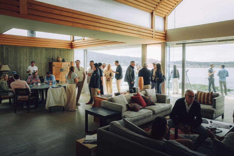 Inside SOC’s Monterey Debut: Convoys Exclusives and Touring's Veloce12 Barchetta Reveal, a group of people socialize indoors and on a patio with large windows overlooking a stunning waterfront view.